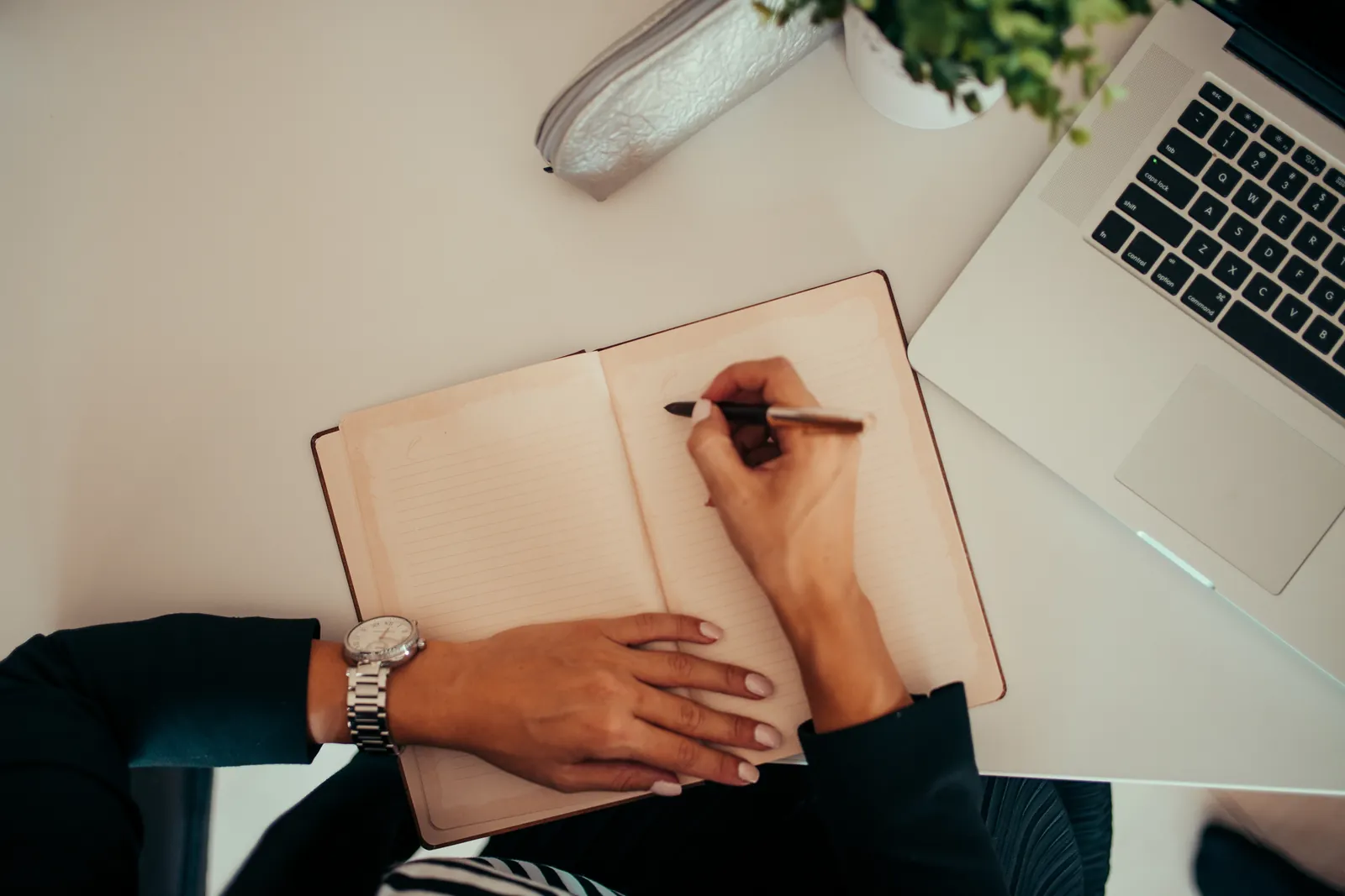 A person writing in a notebook while using a laptop at a modern workspace.