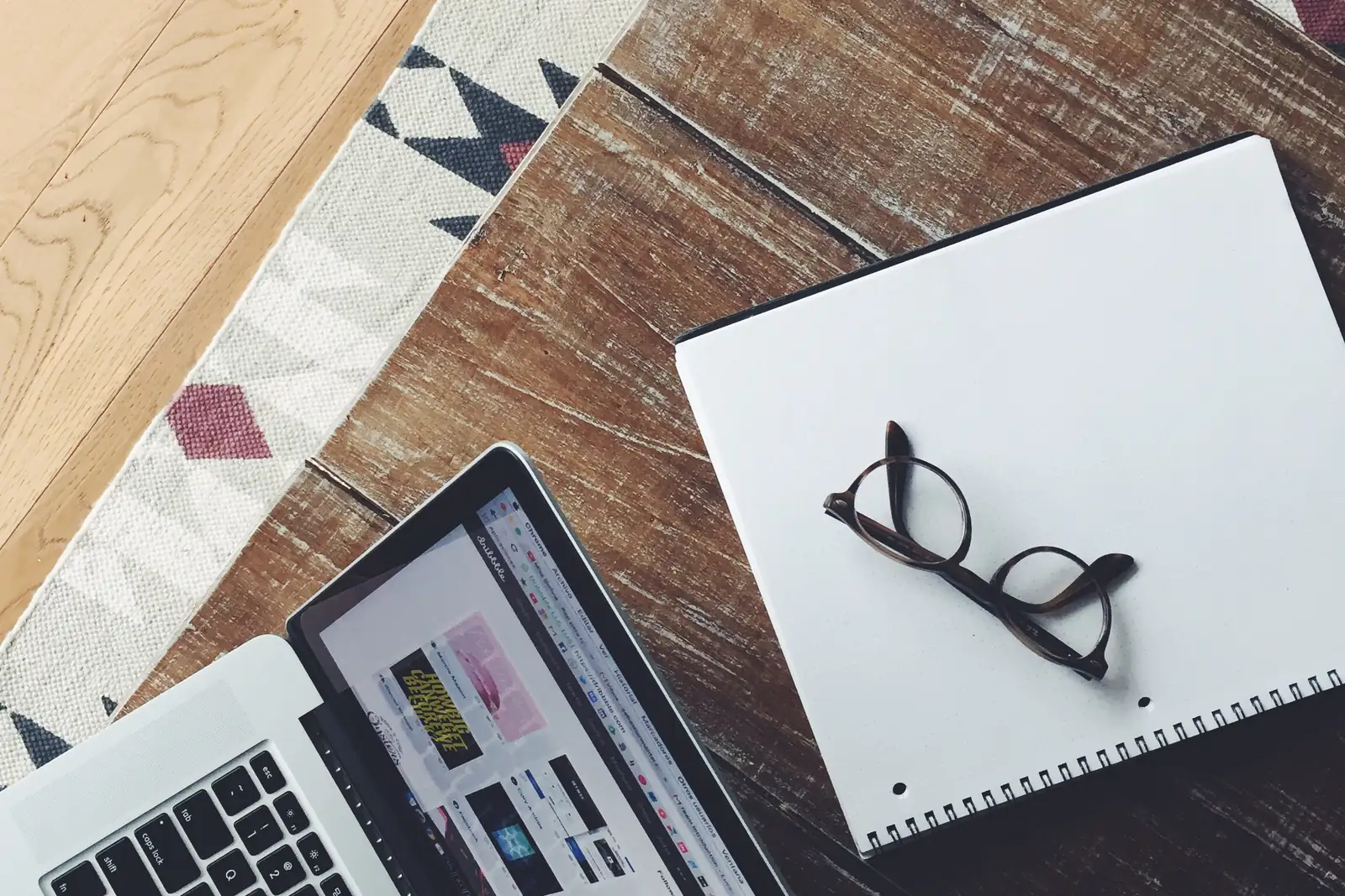 A laptop, notebook, and glasses on a wooden desk.